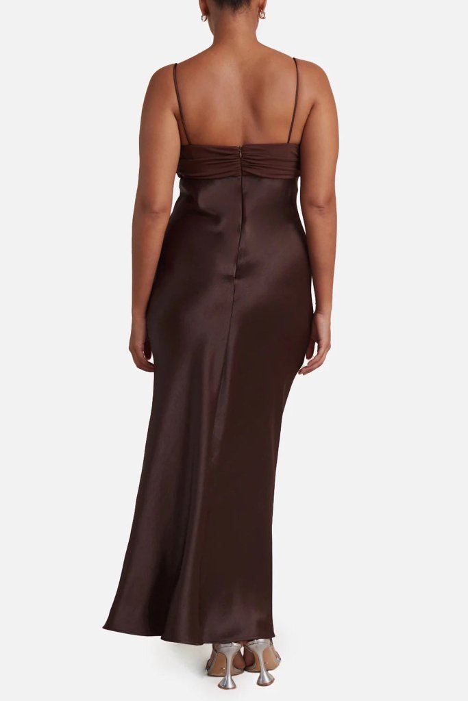 A woman stands with her back to the camera wearing The Wedding Wardrobe's Bec & Bridge Julietta Maxi in Size 10, showcasing a long, dark brown satin fabric with thin straps. The fitted dress elegantly flows to the floor and reveals a high side leg split. Silver high-heeled shoes complete her sophisticated look against the plain white backdrop.