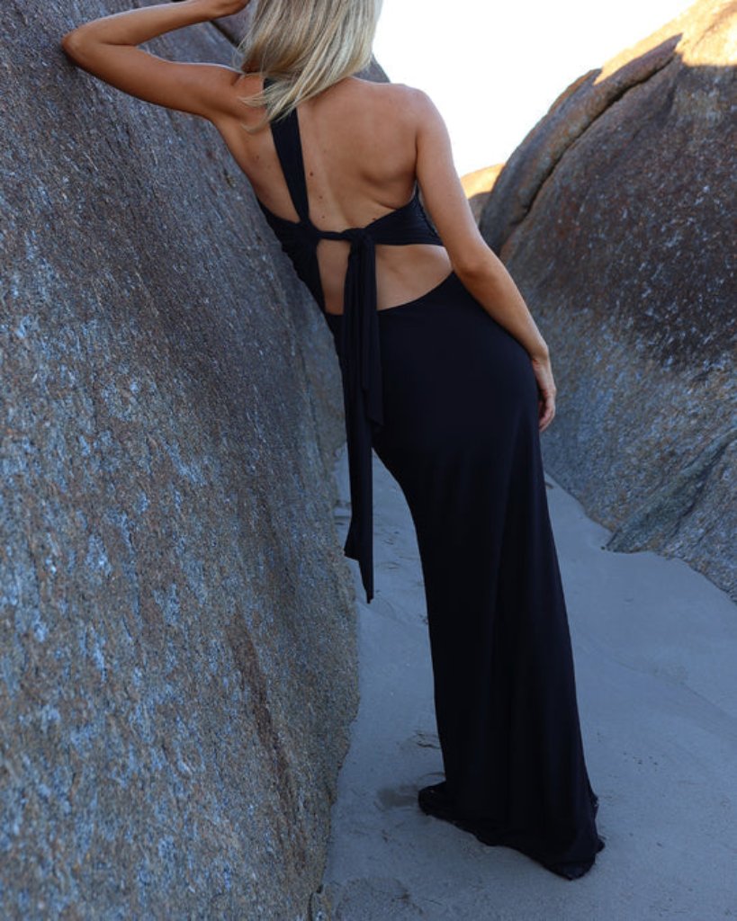 A person with long blonde hair is positioned between large rocks on a sandy path, showcasing the Natalie Rolt Fabienne Gown from The Wedding Wardrobe. The black backless gown, featuring tied straps, elegantly complements the feminine silhouette against the sunlit natural landscape.