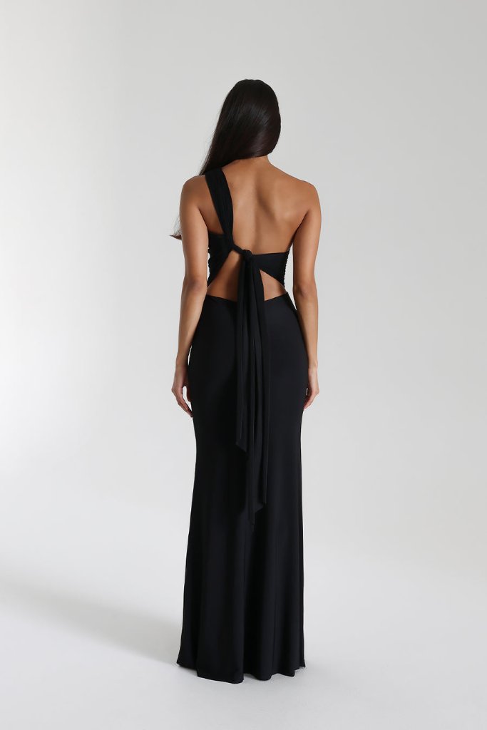 A person with long, dark hair is wearing the elegant Natalie Rolt - Fabienne Gown from The Wedding Wardrobe, in size 10. This stunning black gown features a backless design with a single shoulder strap and sash tie made from Stretch Cavalli fabric. It beautifully highlights their feminine silhouette as they stand against a plain white background.