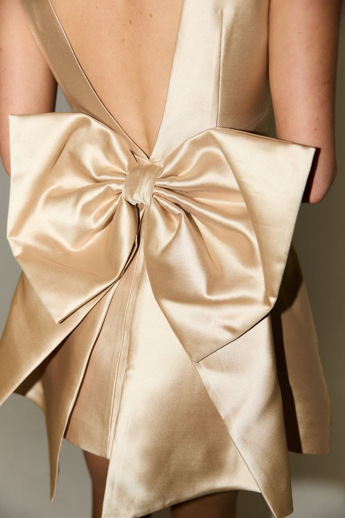 A person wearing "The Ultimate Muse Bow Detail Mini" by The Wedding Wardrobe, a champagne-colored satin shift dress in size 12. It showcases a striking statement bow detailing at the back, complemented by a deep V-back design and a short hemline. The fabric boasts a shiny, smooth texture.
