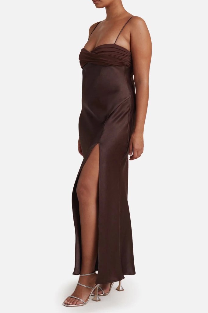 A person wearing the Julietta Maxi by Bec & Bridge, available in size 10 at The Wedding Wardrobe, stands against a white background. This long, sleeveless brown dress features a twisted bandeau and a high side leg split, paired with strappy high-heeled shoes. The dress has a fitted design with a subtle sheen, adding elegance to the ensemble.